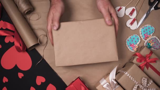 Mãos masculinas sobre a mesa com presentes colocar uma caixa embrulhada em papel artesanal. colocar um grande arco de luz sobre ele. Conceito de dia dos namorados. — Vídeo de Stock