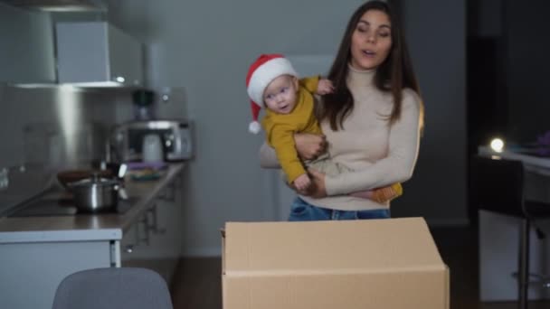 En kvinna med europeiskt utseende håller ett litet barn i sina armar. Jultomtens hatt. Öppna en stor låda — Stockvideo