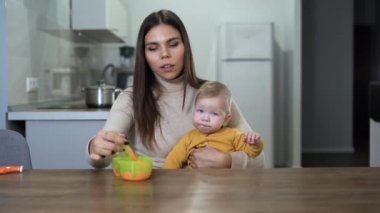 Anne kucağındaki çocuk meyve yiyor, kaşıkla besleniyor. Mutfaktaki beyaz çocuk annesinin yardımıyla bir şeyler atıştırıyor..