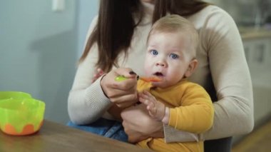 Anne kucağındaki bebek meyve yer, kaşıkla beslenir. Mutfaktaki beyaz çocuk annesinin yardımıyla bir şeyler atıştırıyor..