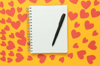 Sarı arkaplandaki büyük boş sarmal not defterinin üst görüntüsü. Bir sürü kırmızı kağıt kalp. Uzayı kopyala Sevgililer Günü konsepti