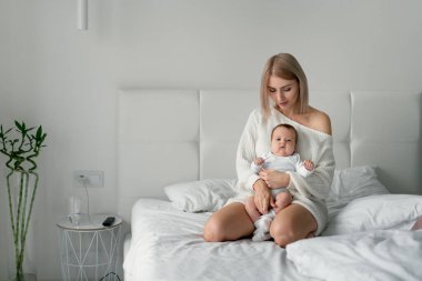 Annem yeni doğmuş bebekle yatakta oturuyor. Kadın bebeği iki eliyle tutuyor..