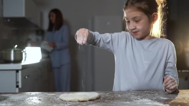 Pizza zu Hause zubereiten, steht ein kleines Mädchen am Tisch und gießt Mehl auf eine rohe Kruste. — Stockvideo
