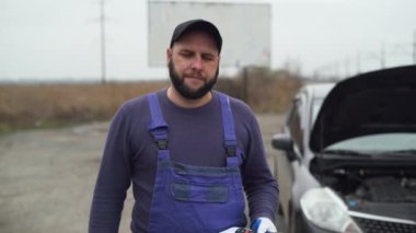 Erkek araba çilingiri bozuk arabanın yanında duruyor. Baş parmağını sallar ve başını sallar..