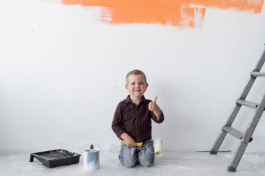 Gülümseyen genç bir çocuk merdivenlerin yanındaki bir odada oturuyor. Elinde baş parmağıyla bir boya silindiri tutuyor. Ev yenileme konsepti.