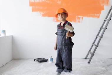 Tulum giymiş ve koruyucu kasklı bir çocuk elinde boya fırçasıyla odada duruyor. Ev yenileme konsepti.