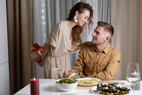 young couple at the table, a woman bent over a man holding a box with a gift in his hands. concept of celebration of valentine's day, anniversary, birthday,