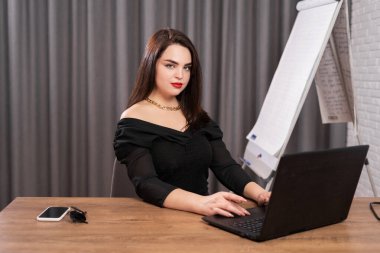 Online sunum. Genç bir kadın bir tablonun yanında oturuyor. Aerodinamik modüllerle gündelik giysiler içinde modern parlak bir odada dizüstü bilgisayarın önünde. Evden çalış.