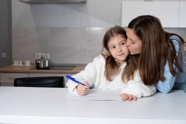 mom and daughter together in the kitchen, the girl draws at the table, her mother kisses her on the cheek. Happy single mother concept. copy space.
