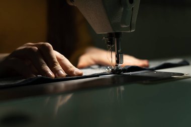 Işıkla aydınlatılan kızın elleri, kumaşı dikiş makinesinin iğnesinin altına yerleştirirken kıyafetleri dikiyor. Dikiş atölyesi konsepti. Metnin için yer.