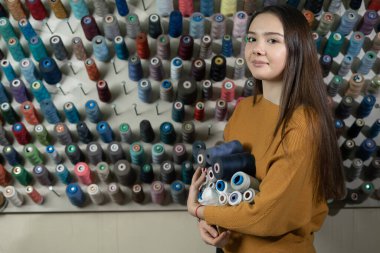 Atölyedeki genç bir terzi kız farklı renklerde ipliklerle bir standın yanında duruyor. Ellerinde çeşitli çürükler tutuyor. Gömlek dikmek için dikiş malzemesi seçimi. Boşluğu kopyala.