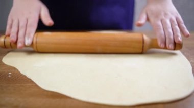 Kadın elleri hamuru ahşap bir yüzeye merdaneyle yakın çekimde yuvarlıyor. Kadın eli masaya hamur serer..