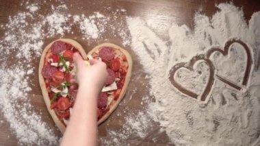 Masanın üzerinde kalp şeklinde büyük bir pizza uzanıyor, bir kadının eli üzerine peynir koyuyor. Kalp şekli, sevgililer günü için akşam yemeği konsepti, yıldönümü, aşkla