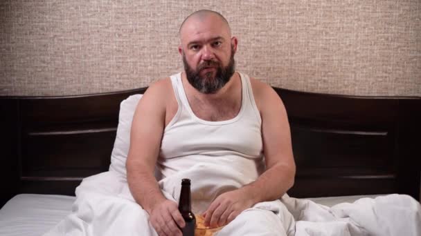 Egy szakállas és nagyra nőtt férfi ül otthon egy ágyon pólóban, sört iszik és chipset eszik. unalmas arckifejezés. A férfiak életkorválságának koncepciója — Stock videók