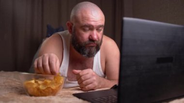 Yetişkin sakallı bir adam yatağa uzanıp film izliyor ve cips yiyor. Çevrimiçi sinema kavramı