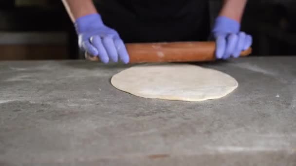 Um chef italiano esculpe a massa em uma superfície com farinhas e a enrola com um rolo na cozinha tradicional da pizzaria. close-up de mãos em luvas — Vídeo de Stock