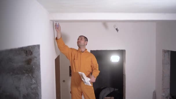 Καυκάσιος εργάτης σοβατίζει το ταβάνι, φορώντας πορτοκαλί στολή. Σχέδιο ανακαίνισης σπιτιού — Αρχείο Βίντεο