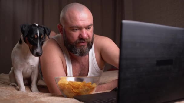 Un uomo barbuto adulto in abiti da casa è sdraiato sul letto e guardare un film e mangiare patatine fritte. Il prossimo e 'Cane Jack Russell. Concetto di cinema online — Video Stock