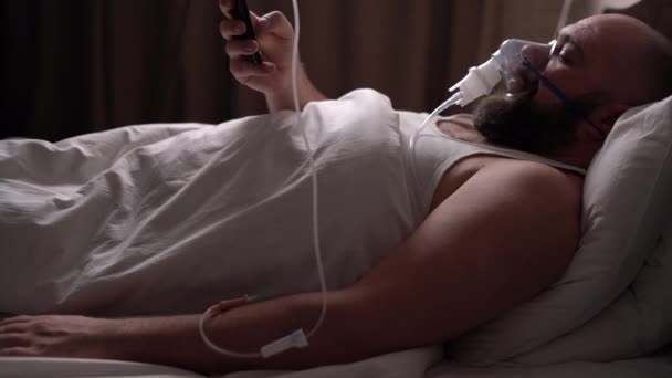 人工呼吸器とドロッパーに接続されたベッドの上に男が横たわっている。スマートフォンにメッセージを送ることです. — ストック動画