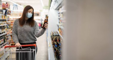 Koruyucu maskeli bir kadın, marketin yanındaki raflarda elinde alışveriş arabasıyla durup elindeki cam şişe biraya bakar..