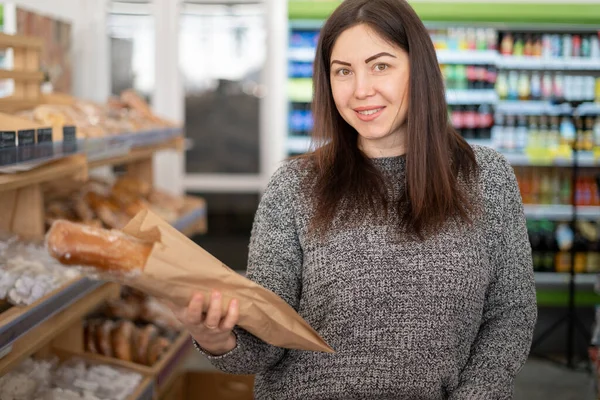 Брюнетка стоїть біля полиць у пекарні, тримаючи пшеничний хліб у паперовій сумці в руках. Shopper жінка дивитися . — стокове фото