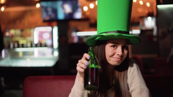 Mladá žena slaví den svatého Patricka v baru během večírku. Velký zelený klobouk sedí u stolu a pije pivo z láhve na hlavě. — Stock video