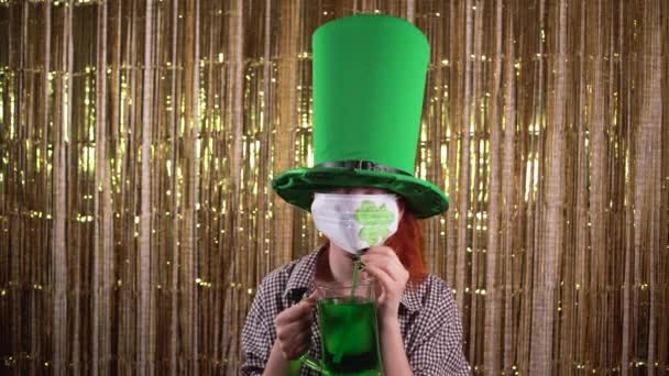 Giovane donna che celebra giorno st patricks, vestito con un costume folletto con una maschera protettiva sul viso, bere birra verde attraverso una cannuccia. — Video Stock