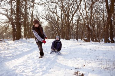 Çocuklar kışın parkta kızak kullanırdı. Neşeli bir kız kızağı sürüklüyor, bir oğlan onların üstüne oturuyor..