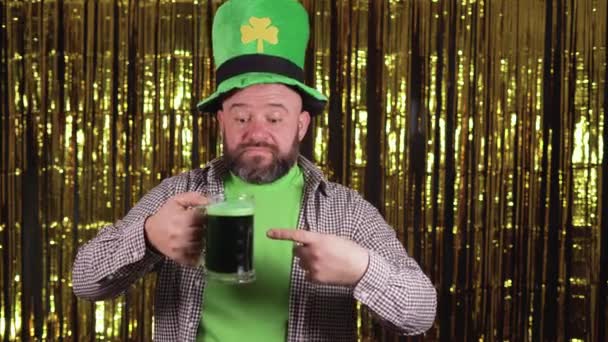 Un uomo caucasico barbuto tiene in mano un bicchiere di birra verde e gli punta un dito contro. Santo patricks giorno celebrazione concetto. — Video Stock