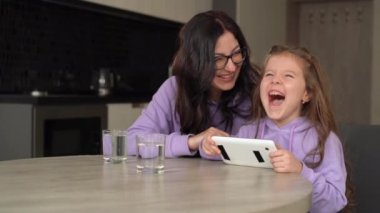 Sarışın, kıvırcık saçlı güzel bir kız ve gözlüklü esmer annesi mutfakta bir masada oturmuş tablet üzerinde çizgi film izliyorlar..
