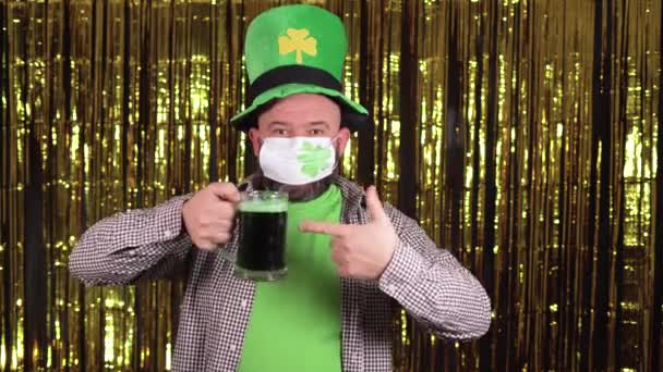 顔に医療マスクをした男がパティックの日を祝う。指の先に緑のビールのグラスを持っていて. — ストック動画