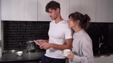 Genç çift kahvaltıda mutfakta tablet tablet kullanıyor. Elinde kahve tutan ve interneti dijital tabletle gezen sevimli ev hanımı.