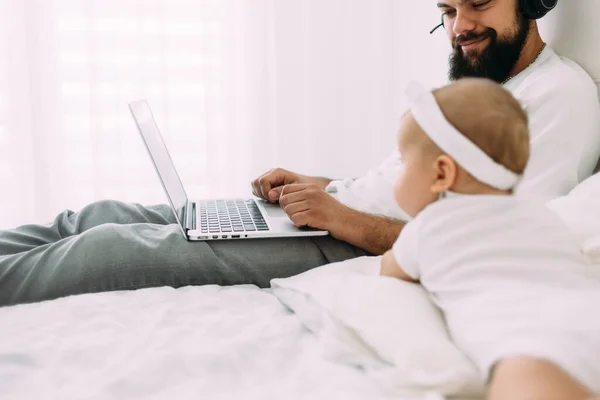 Dad works remotely with headphones, and his daughter lies next to him on the bed. Fatherhood and childhood. Family time and earnings.