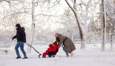 Babam, üç yaşındaki kızını ağaçların arasındaki kış parkında karda kızakla gezdirirken, annem arkadan itiyor. Kışın aile tatili. Boşluğu kopyala.