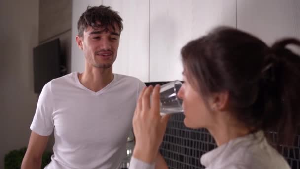 Casal jovem no fundo da cozinha. uma mulher segura um copo de água limpa em suas mãos e bebe. — Vídeo de Stock