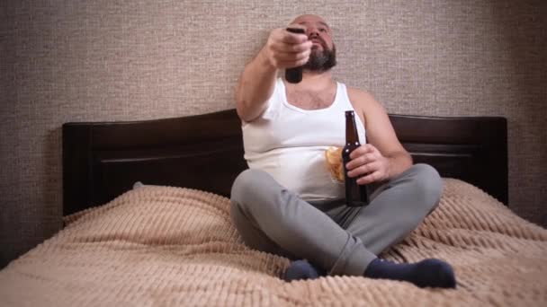성인 백인 남자 평상복을 입고 침대에 앉아 있습니다. 손에 맥주와 감자칩을 쥐고 TV 채널을 리모 콘으로 전환 합니다. — 비디오