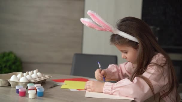一个长着小兔子耳朵的可爱女孩正准备迎接复活节，她坐在桌旁，在纸上画着一只野兔. — 图库视频影像