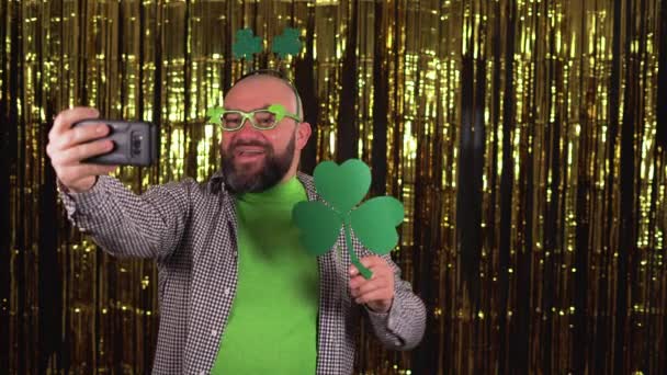Een bebaarde Europese man viert St Patricks Day, shamrock hoorns op zijn hoofd en een groene bril, maakt een videogesprek. — Stockvideo