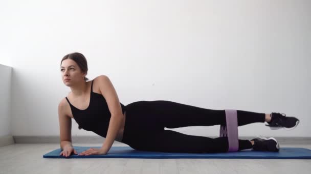 A girl at home using elastic rubber trains her lower muscles lying on the mat, a determined athlete reaching fitness goals and losing weight during a hard workout — Stock Video