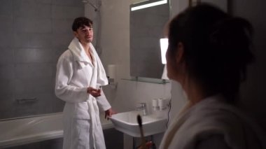 Genç bir Avrupalı banyoda bornozlu ve dişlerini fırçalıyor. Kız arkadaşı diş minesiyle ilgilenen adamı izliyor..