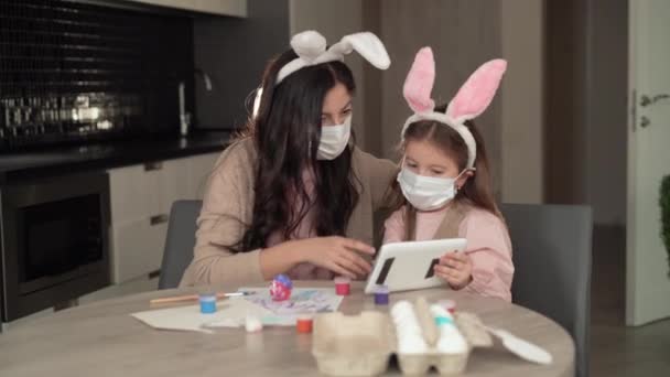 행복 한 부활절. 한 쾌활 한 가족 이 부활절 연휴를 준비하고 있다. 의료용 마스크를 쓴 어머니와 딸은 부엌에 있는 집에 앉아 있습니다. 태블릿을 통해 비디오 통화를 한다 — 비디오