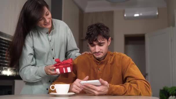 Jeune femme en apparence donne un cadeau à un homme. Le gars est assis avec une tablette dans ses mains et la main refuse un cadeau. — Video