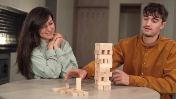 Młoda para siedzi w domu podczas kwarantanny i gra w grę planszową Jenga. Mężczyzna ubrany w koszulę w koncentracji wyciąga jeden drewniany prostokąt. — Wideo stockowe