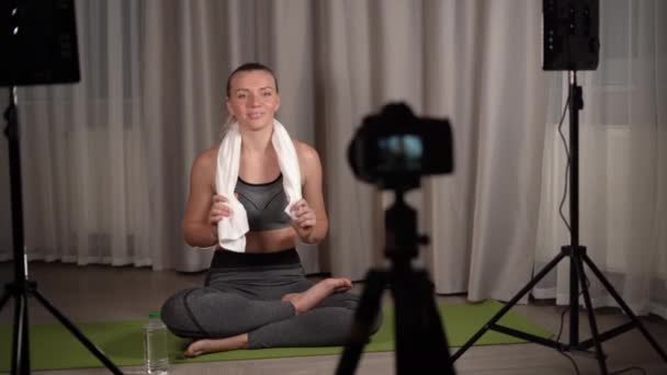 Concepto de fitness, deportes y vlogging - mujer o blogger con cámara en la grabación de trípode lección de yoga en línea. charla alegremente y limpia el sudor de su frente. — Vídeo de stock