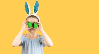 Happy Easter, kids. A girl in rabbit ears on her head holds two decorated eggs in her hands and closes her eyes with them. Baner, copy space