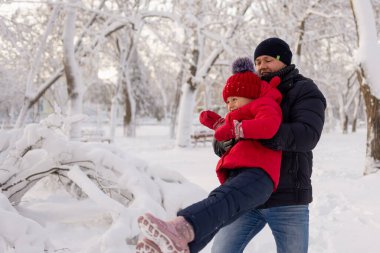 Karlı bir kış parkında, mutlu genç bir adam kızını kırmızı bir ceketle kollarına aldı ve onu döndürdü. Kışın seyahat ve aile tatili..