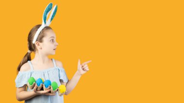 Mutlu Paskalyalar, çocuklar. Tavşan kulaklı bir kız elinde mavi ve sarı renkli yumurtalarla dolu bir tepsi tutuyor. Yüzünde şaşkınlık hissi var. İşaret parmağı metin için boş bir alanı gösterir.