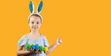 Happy Easter, kids. A girl with bunny ears on her head holds a tray of blue and yellow eggs. The index finger points to an empty space for text.