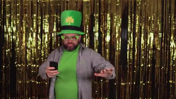 Mužský skřítek oblečený ve velkém zeleném klobouku slaví den svatého Patricka, tančí s telefonem na žlutém pozadí, přibližuje se ke kameře a je překvapen. — Stock video