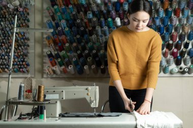 Beyaz bir terzi çalışma masasında duruyor ve beyaz kumaşı makasla kesiyor. Atölyedeki terzi sevdiği şeyi yapıyor. Metin için yer.